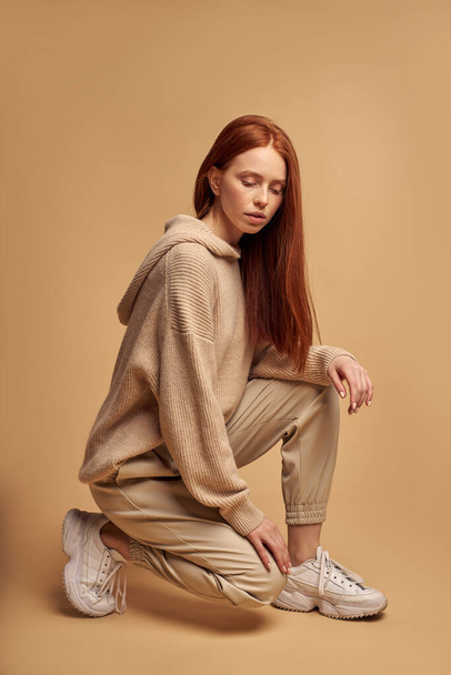 Young redhead female in beige casual wear sitting on floor in studio isolated - Foto, Imagem