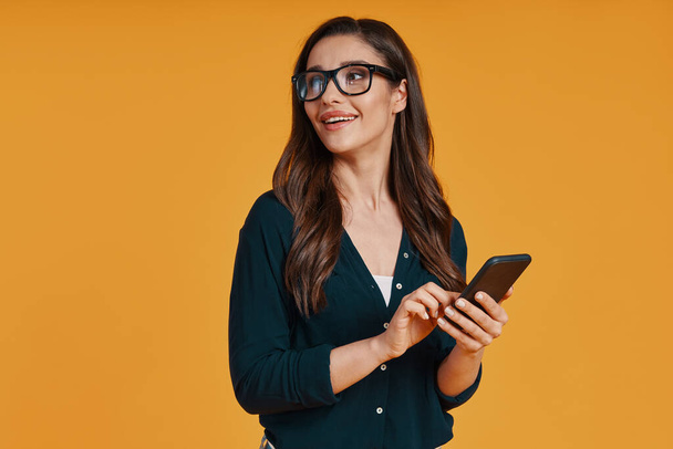 Beautiful young woman in casual clothing smiling and using smart phone while standing against yellow background - Φωτογραφία, εικόνα
