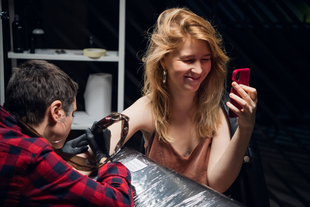 Девушка фотографируется для своих социальных сетей, в то время как профессиональный татуировщик кладет татуировку на руку, используя палку и метод тычка. - Фото, изображение