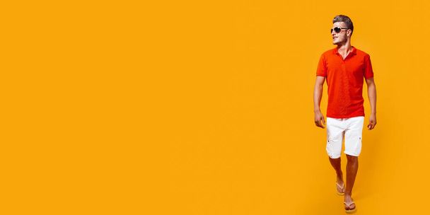 Full length portrait of a handsome man in shorts and red shirt walking and looking sideway isolated on yellow background. Summertime concept - Foto, Bild