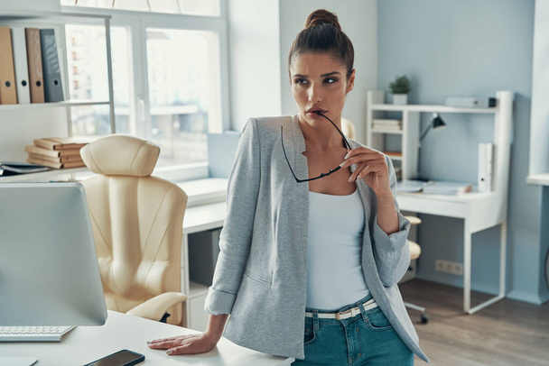 Thoughtful young woman in smart casual looking away and holding eyewear while standing in the office - Фото, изображение