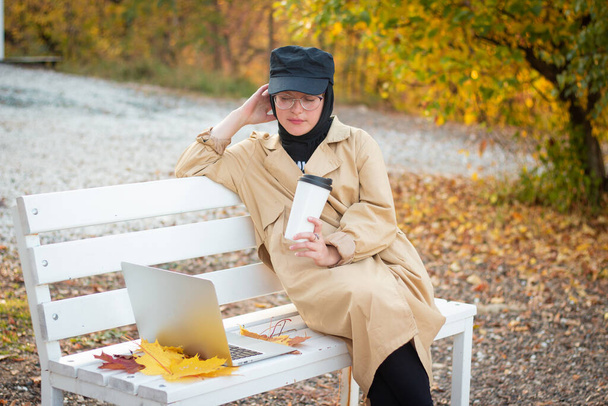 A Muslim woman in a hijab in a beige trench coat sits on a bench with a cup of coffee and a laptop in the park. Muslim business woman. Student. Glasses for sight. - Foto, afbeelding