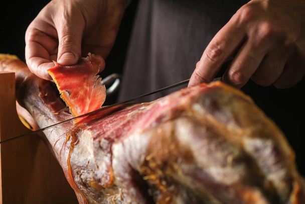 Dry Spanish ham, Jamon Serrano, Italian Prosciutto Crudo or Parma ham, whole leg on black background.A jamon slicer cuts a slice of meat.Slice of ham in a man's hand. - Photo, Image