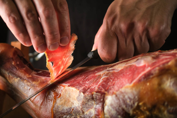 Dry Spanish ham, Jamon Serrano, Italian Prosciutto Crudo or Parma ham, whole leg on black background.A jamon slicer cuts a slice of meat.Slice of ham in a man's hand. - Fotó, kép