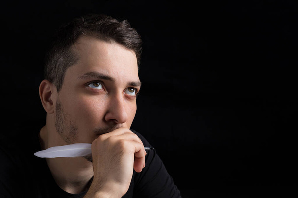 A young handsome man in black clothes with a goose feather looks pensively and romantically up, close-up, on a black background in a low key. Copyspace for text - Photo, Image