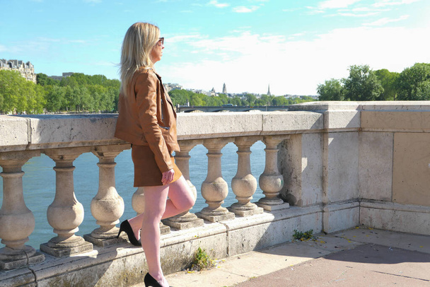 Mooie blonde vrouw in rok met stiletto hakken, staande op een historische stenen brug, met uitzicht op de Eiffeltoren in de stad. Achtergrond vervaagd vrijwillig. - Foto, afbeelding