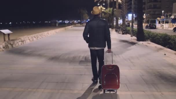 Un turista con una maleta roja vino a descansar tarde por la noche, camina por el terraplén - Metraje, vídeo