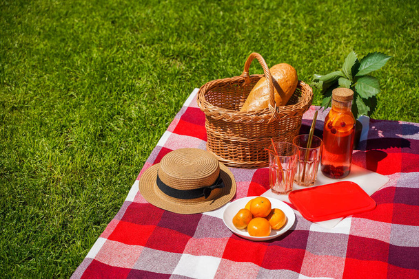 Piknik nurmikolla, punainen ruudullinen huopa, pajukori, appelsiineista ja marjoista valmistettu luonnollinen juoma, leipä. Herkullinen välipala ulkona kesällä - Valokuva, kuva