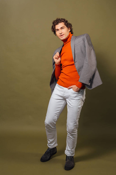 Full length portrait of a tall handsome man dressed in orange turtleneck, grey jacket and white jeans posing on the green background - Photo, image