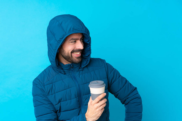 Man wearing winter jacket and holding a takeaway coffee over isolated blue background with arms crossed and happy - Foto, imagen