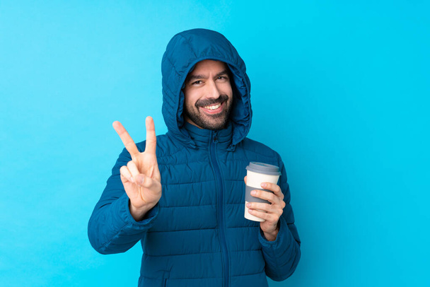 Man wearing winter jacket and holding a takeaway coffee over isolated blue background smiling and showing victory sign - Foto, imagen