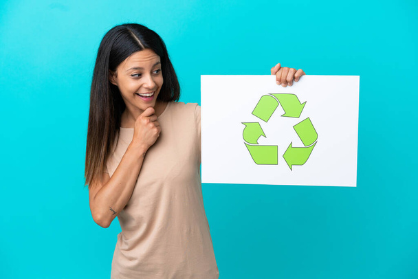 Mujer joven sobre fondo aislado sosteniendo una pancarta con icono de reciclaje - Foto, imagen
