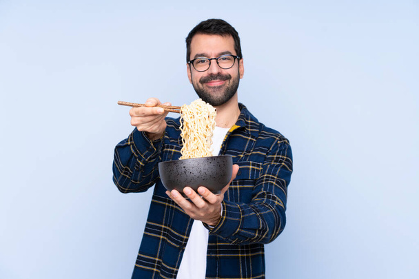 Young caucasian man over blue background holding a bowl of noodles with chopsticks and offering it - Foto, Imagen