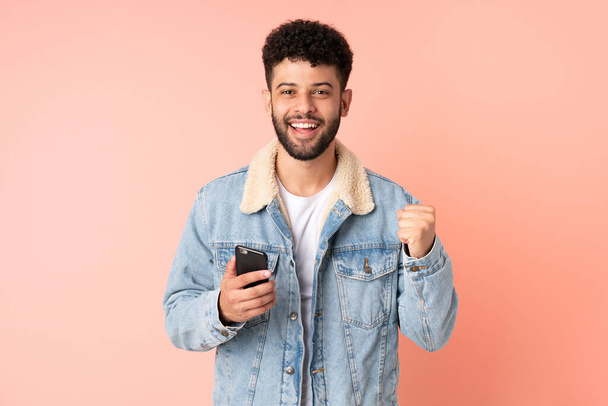 Young Moroccan man using mobile phone isolated on pink background celebrating a victory in winner position - Fotografie, Obrázek