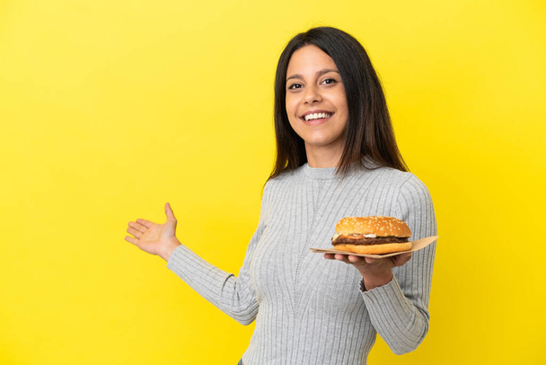 Sarı arka planda izole edilmiş hamburgerini tutan genç beyaz kadın davet etmek için elini yana uzatıyor. - Fotoğraf, Görsel