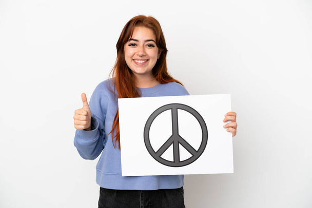 Young redhead woman isolated on white background holding a placard with peace symbol with thumb up - 写真・画像