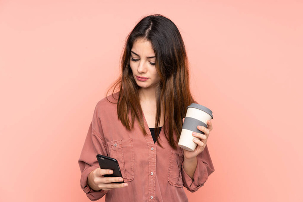 jonge Kaukasische vrouw geïsoleerd op roze achtergrond houden koffie mee te nemen en een mobiele - Foto, afbeelding