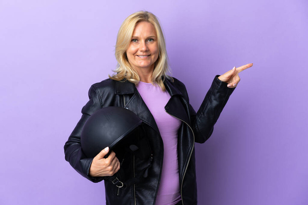 Middle age woman holding a motorcycle helmet isolated on purple background pointing finger to the side - Fotoğraf, Görsel