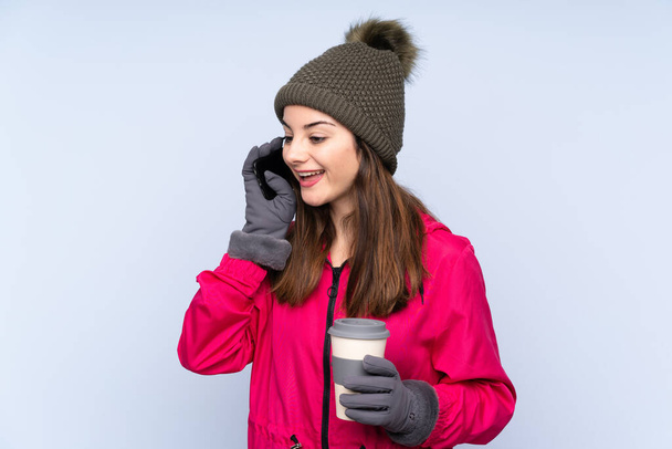 Young girl with winter hat isolated on blue background keeping a conversation with the mobile phone - Photo, image