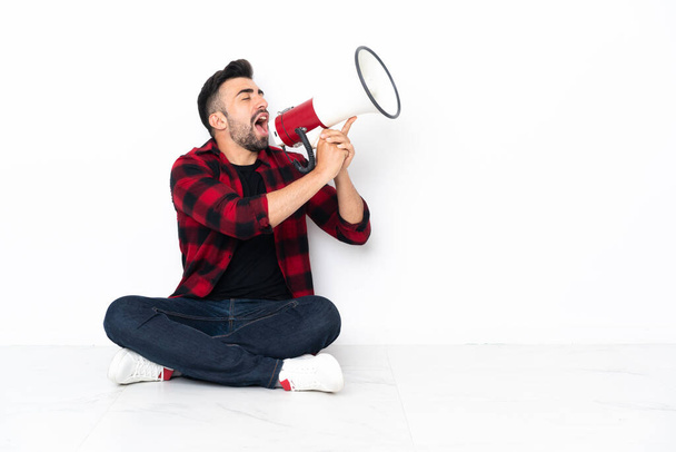 Young handsome man sitting on the floor shouting through a megaphone - Photo, Image