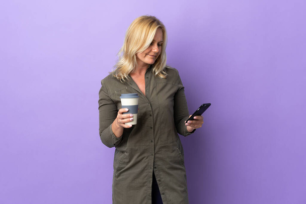 Middle age Lithuanian woman isolated on purple background holding coffee to take away and a mobile - 写真・画像