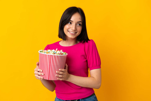 Young caucasian woman isolated on yellow background holding a big bucket of popcorns - Photo, Image