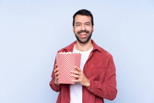 Young caucasian man over blue background holding a big bucket of popcorns - Fotoğraf, Görsel