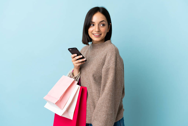 Giovane donna caucasica isolata su sfondo blu con borse della spesa e un telefono cellulare - Foto, immagini