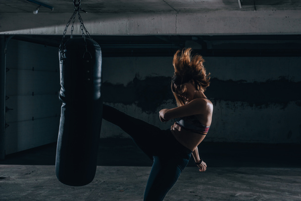 Genç siluet kadın garajda boks çantasıyla yüksek tekme pratiği yapıyor.. - Fotoğraf, Görsel