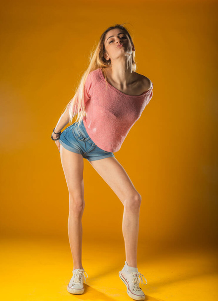 Sportowa śliczna dziewczyna w dżinsowych spodenkach, tańcząca z przyjemnością i uśmiechnięta. Indoor portret oszałamiający model kobieta z vintage fryzury machanie rękami na żółtym tle. - Zdjęcie, obraz