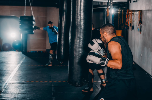Férfi bokszoló edzés boxzsákkal a sötét sportcsarnokban. Férfi bokszoló, mint testmozgás a nagy harcra. Boxer találat boxzsák - Fotó, kép