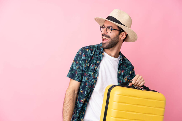 Młody mężczyzna kaukaski na odizolowanym tle na wakacjach z walizką podróżną i kapeluszem - Zdjęcie, obraz