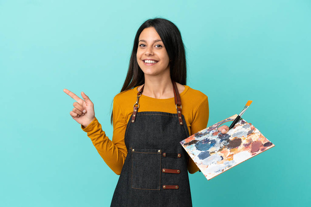 Mladý umělec běloška žena drží paletu izolované na modrém pozadí ukazující prstem na stranu - Fotografie, Obrázek