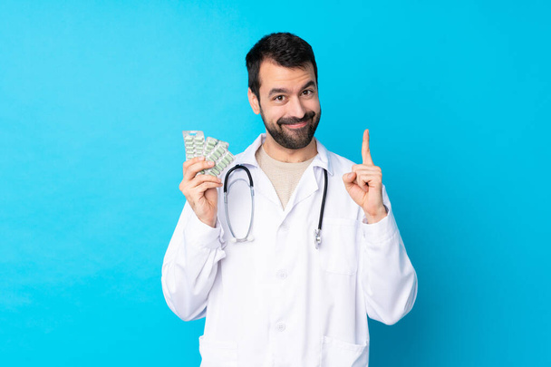 Young caucasian man over isolated background wearing a doctor gown and holding pills - Photo, image