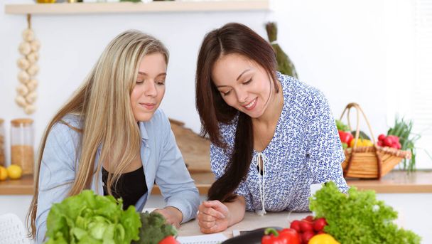 Two women friends choosing the recipe for a delicious meal while sitting at the table in the kitchen. Tablet pc is the best cookbook. - Φωτογραφία, εικόνα