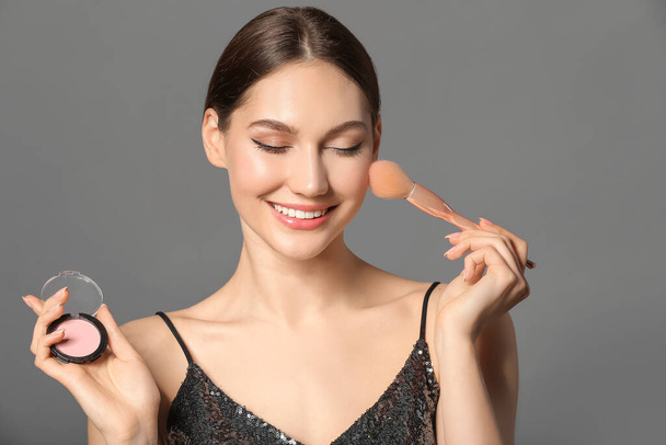 Mooie jonge vrouw met make-up borstel en blozen op grijze achtergrond - Foto, afbeelding