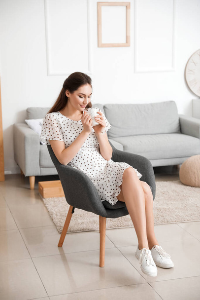 Νεαρή γυναίκα πίνει καφέ ενώ κάθεται στην πολυθρόνα στο σπίτι - Φωτογραφία, εικόνα