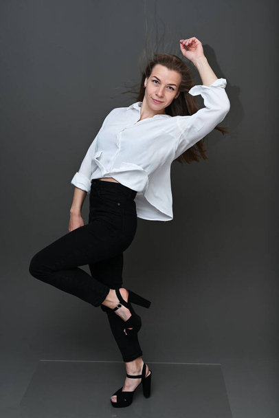 Portrait of a Caucasian girl with fluttering hair in the wind in black trousers and a white blouse with a smile on a gray background in full growth. Studio photo in high resolution. - Фото, изображение
