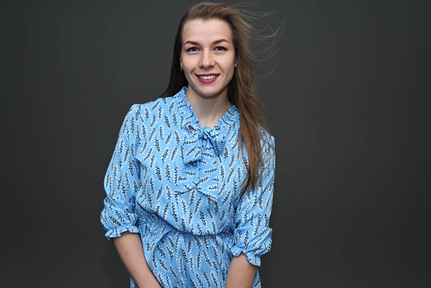 Portrait of a Caucasian girl with fluttering hair in the wind in a blue dress with a smile on a gray background. Studio photo in high resolution. - Photo, image