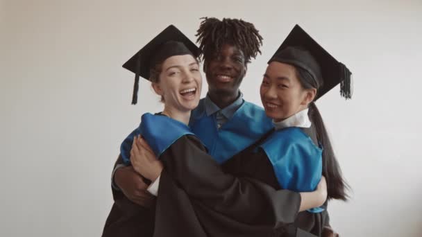 Portrait ralenti moyen de trois étudiants des cycles supérieurs caucasiens, asiatiques et afro-américains en casquettes et robes souriant à la caméra s'embrassant debout sur fond blanc - Séquence, vidéo