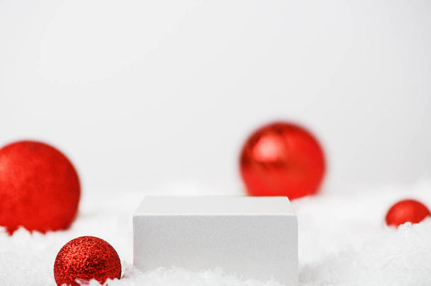 Bílý kostkový podstavec na sněhu na zimním slavnostním bílém pozadí. Lehká vánoční scéna s pódiem. Výstava kosmetiky a kosmetických výrobků. Stánek s novoroční výzdobou s červenými koulemi. - Fotografie, Obrázek