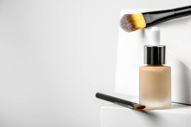Beige foundation on white stand with makeup brushes. BB cream, nude concealer, perfect skin tone. Beauty liquid skincare fluid. Female cosmetic accessory with geometric light background. Copy space - Photo, Image
