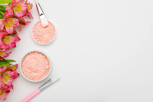 Natural facial mask in jars and with brushes on white background with flowers. Pink alginate clay mask with kaolin and collagen, lifting effect. Cleansing cosmetic beauty product.Peeling,facial scrub. - Fotografie, Obrázek