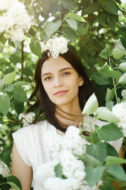 Portrait of a beautiful young Caucasian brunette girl on a background of white blooming lilac on a warm sunny day. Flowers in your hair. The concept of women's beauty and health - Fotoğraf, Görsel
