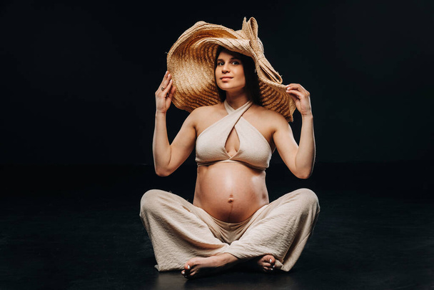 a pregnant woman in a straw hat sits on the floor in beige clothes in a studio on a black background. - Fotoğraf, Görsel