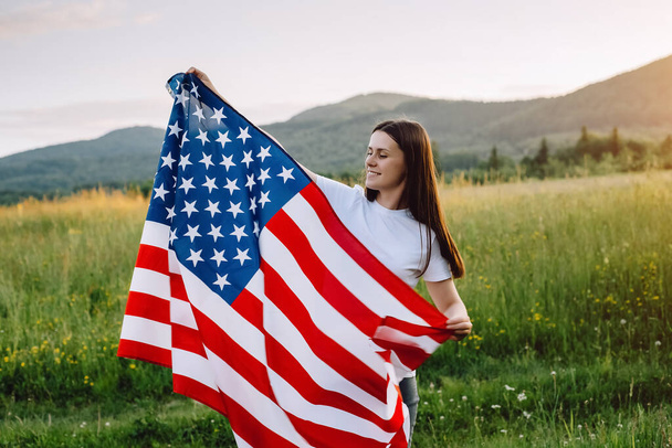 Happy smiling young brunette woman with american flag standing in middle meadow on background mountain during sunset. 4th of July. Independence Day. Patriotic holiday. USA flag fluttering in wind - Fotoğraf, Görsel