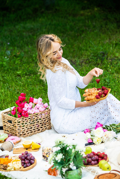 Mooie vrouw rustend in het park zittend op een picknickdeken met fruit en wijn.  - Foto, afbeelding