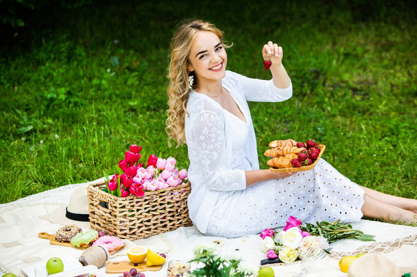 Όμορφη γυναίκα αναπαύεται στο πάρκο κάθεται σε μια κουβέρτα πικνίκ με φρούτα και κρασί.  - Φωτογραφία, εικόνα
