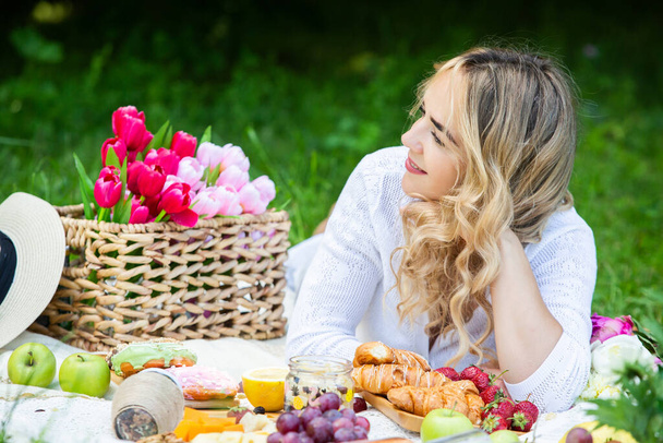 Piękna kobieta odpoczywająca w parku, siedząca na kocu piknikowym z owocami i winem.  - Zdjęcie, obraz
