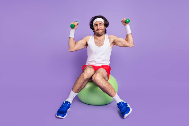 Full size of young athlete funky man in glasses sit ball lifting little dumbbells trying hard isolated on violet color background - Photo, Image